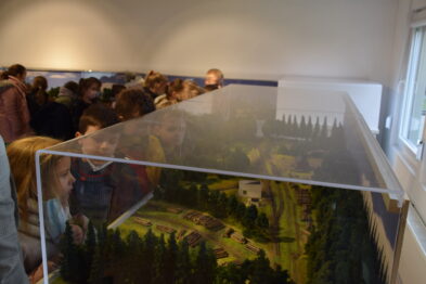 Grupa osób stoi wokół dioramy przedstawiającej scenerię kolejową. Wśród obserwujących dominują dzieci, które uważnie przyglądają się modeleowi. Diorama jest zabezpieczona pleksi, a pomieszczenie oświetlone sztucznym światłem.