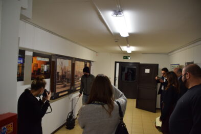 Grupa ludzi ogląda zdjęcia o tematyce kolejowej zawieszone na ścianach białego korytarza w galerii. Kilku odwiedzających robi zdjęcia ekspozycji, która obejmuje kadry z pociągami i tory kolejowe. Jest to wernisaż, gdzie uczestnicy stoją pod światłami sufitowymi, co tworzy przyjemną atmosferę spotkania z fotografią.