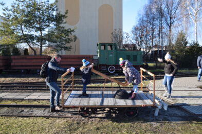 Grupa osób stoi i siedzi na drewnianym platformowym wagoniku kolejowym, który jest umieszczony na wąskotorowych szynach. W tle widnieje zielona lokomotywa oraz wagony towarowe. Uczestnicy wydarzenia są ubrani w zimowe kurtki, a jedna osoba ma na głowie karnawałowy kapelusz.