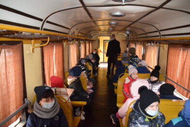 Grupa dzieci siedzi na ławkach wewnątrz wagonu kolejowego, każde z nich ma na twarzy maseczkę ochronną. Pasażerowie są ubrani w zimowe odzieże takie jak czapki, szaliki i kurtki. Na środkowym przejściu wagonu stoi dorosły opiekun ubrany w ciemny płaszcz, obserwujący uczestników wycieczki.