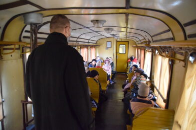 Wagon wąskotorowego pociągu jest wypełniony ludźmi; na drewnianych ławkach siedzą dzieci i kilka osób dorosłych. Na środku przejścia stoi mężczyzna plecami do kamery, obserwujący uczestników zajęć. Wnętrze jest jasne, z elementami drewnianymi oraz zaokrąglonym sufitem.