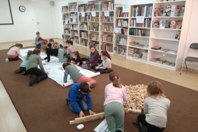 Grupa dzieci siedzi na podłodze i angażuje się w różnorodne aktywności, jedna osoba tworzy konstrukcje z drewnianych klocków. Inne dzieci rysują lub malują na dużych arkuszach papieru rozłożonych wokół siebie. W tle widoczne są półki z książkami oraz plakaty na ścianie.