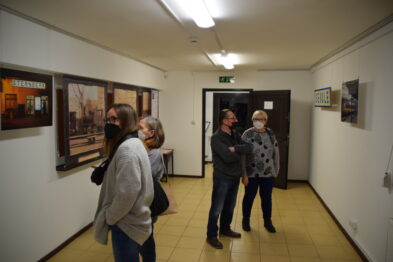 Grupa osób ogląda fotografie o tematyce kolejnictwa zawieszone na ścianach jasnego korytarza. Widać trzy kobiety i mężczyznę stojących w galerii - dwie z kobiet są w rozmowie, a pozostałe osoby skupiają swój wzrok na jednym z eksponatów. Przestrzeń jest dobrze oświetlona, co pozwala na wyraźne dostrzeżenie detali prezentowanych prac.
