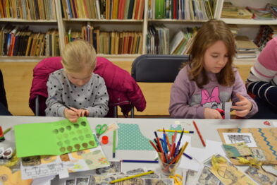 Dwie dziewczynki siedzą przy stole i tworzą rękodzieła lub pracę plastyczną. Na stole rozłożone są arkusze papieru, kolorowe kredki oraz inne materiały plastyczne takie jak naklejki z motywami kolejowymi. Dzieci są skupione na swoich projektach, a otoczenie wydaje się być przestrzenią edukacyjną lub warsztatową.
