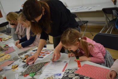 Dzieci wraz z dorosłą osobą biorą udział w zajęciach plastycznych przy dużym stole, gdzie rozłożone są kolorowe papiery i przybory do rysowania. Osoba dorosła pomaga jednemu z dzieci w pracach manualnych. Wokół panuje atmosfera skupienia i twórczej aktywności.