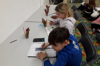 Dzieci siedzą przy białym stole i rysują kredkami, skoncentrowane na swoich zadaniach. Stół jest pokryty kolorowymi kredkami i rozłożonymi kartkami papieru. W tle widoczne są inne dzieci, również zajęte rysowaniem.
