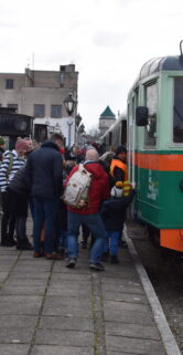 Na peronie grupa osób w kurtkach stoi między dwoma pociągami: z lewej strony parowozem, a z prawej kolejowym pojazdem motorowym. Peron pokryty jest kostką brukową, a w tle widoczne są zabudowania miejskie. Osoby skupiają się wokół parowozu, co sugeruje zainteresowanie ekspozycją kolejową.