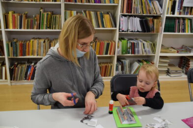 Kobieta i dziecko siedzą przy stole w pomieszczeniu z półkami pełnymi książek. Obydwoje są skupieni na pracy z papierowymi elementami, które leżą przed nimi na stole. W tle widoczne są liczne półki z ułożonymi książkami oraz inne materiały.
