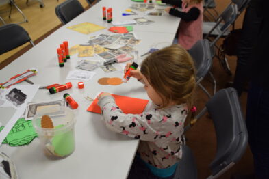 Mała dziewczynka w kolorowej bluzie siedzi przy stole i skupia się na robieniu rękodzieła; na stole rozłożone są przybory plastyczne i kolorowe kartki. Dziecko trzyma w ręce czerwony przedmiot, być może jest to farba lub klej. W tle nieostro można zauważyć innych uczestników zajęć kreatywnych.