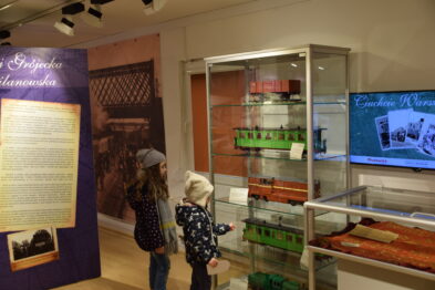 Dwoje dzieci stoi przed szklaną wystawką z modelami kolejowymi, skupiając uwagę na zielonych lokomotywach wystawionych na półkach. Na ścianie obok znajduje się monitor prezentujący slajdy oraz tablice informacyjne z tekstami i zdjęciami. W pomieszczeniu widoczne są także inne eksponaty kolejowe i plakaty.