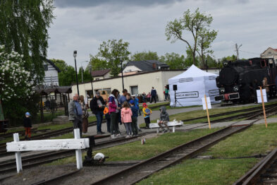 Grupa osób stoi na peronie obok torów kolejowych, gdzie widoczna jest zabytkowa lokomotywa parowa. Na pierwszym planie znajduje się miniatura lokomotywy, którą oglądają dzieci. W tle zauważalne są namioty informacyjne i budynki, które dopełniają organizację wydarzenia.
