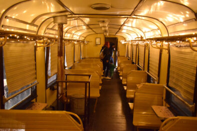 Stare, drewniane siedzenia z metalowymi podstawami układają się w rzędy wewnątrz historycznego wagonu kolejowego zapewniając pasażerom miejsce do siedzenia. Oświetlenie sufitowe dodaje wnętrzu ciepły żółty blask, podkreślając kolorystykę i fakturę drewna. Osoby zwiedzające spacerują wzdłuż przejścia między siedzeniami, oglądając z zaciekawieniem wnętrze wagonu.