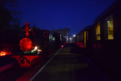 Lokomotywa parowa oświetlona czerwonym światłem stoi na torach obok peronu; po prawej stronie widać szereg wagonów. Jest wieczór lub noc, o czym świadczy ciemne niebo w tle. Atmosfera jest uroczysta i pasująca do nocnego wydarzenia muzealnego.