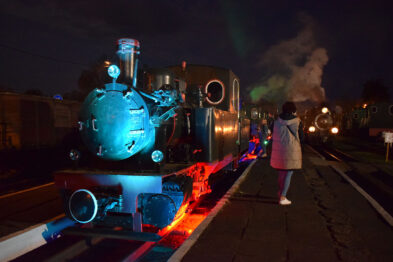 Stara parowa lokomotywa wąskotorowa oświetlona jest kolorowymi światłami podczas nocnej ekspozycji. Dym wydobywa się z jej komina, a tory kolejowe rozciągają się na pierwszym planie obrazu. Przed parowozem stoi osoba w jasnym płaszczu, która obserwuje maszynę.