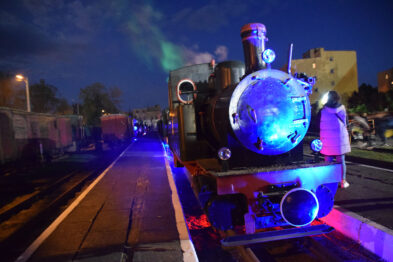 Klasyczna parowa lokomotywa wąskotorowa jest podświetlona kolorowym światłem podczas nocnego wydarzenia. Na pierwszym planie widoczna jest okrągła lampa lokomotywy świecąca na biało, z odbłyśnikiem w kolorze niebieskim. Tłum ludzi zwiedza muzealne ekspozycje, a w tle rozpościera się niebieskie niebo przetykane światłami.