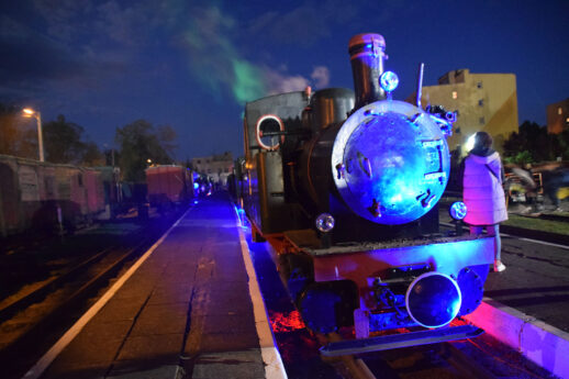 Klasyczna parowa lokomotywa wąskotorowa jest podświetlona kolorowym światłem podczas nocnego wydarzenia. Na pierwszym planie widoczna jest okrągła lampa lokomotywy świecąca na biało, z odbłyśnikiem w kolorze niebieskim. Tłum ludzi zwiedza muzealne ekspozycje, a w tle rozpościera się niebieskie niebo przetykane światłami.