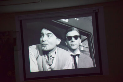Wyświetlany jest czarno-biały film na ekranie projekcyjnym, przedstawiający dwóch mężczyzn siedzących obok siebie. Mężczyzna po lewej stronie ma kapelusz i patrzy w lewo z otwartymi ustami, natomiast mężczyzna po prawej nosi ciemne okulary i patrzy prosto. Otoczenie wskazuje, że znajdują się wewnątrz pojazdu, prawdopodobnie pociągu.