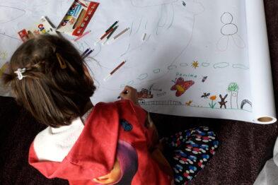 Dziewczynka w czerwonej bluzie siedzi na podłodze i koloruje duży arkusz papieru z rysunkami kolejowymi. Na arkuszu widoczne są rysunki pociągów, torów i różnych elementów związanych z koleją. Obok niej leży zestaw kolorowych kredek.