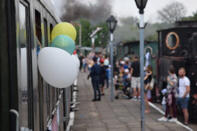 Kolorowe balony są przyczepione do uchwytu na zewnątrz wagonu kolejowego. Grupa osób czeka na peronie, a w tle widać starą parową lokomotywę. Scena odbywa się na terenie muzeum kolejnictwa, atmosfera wydarzenia jest świąteczna i rodzinna.