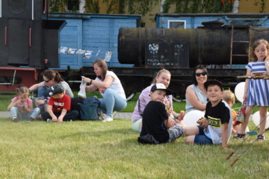 Grupa dzieci siedzi na trawie z dorosłymi podczas wydarzenia w muzeum kolejnictwa. W tle widać stare czarne parowozy. Osoby uczestniczą w plenerowej aktywności na zielonej przestrzeni obok historycznego taboru.