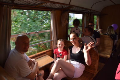 Rodzina podróżuje pociągiem, siedząc na drewnianych ławkach z plecionymi oparciami; widać uśmiechniętą dziewczynkę, kobietę i mężczyznę machających do kamery. Wagon oświetlony jest naturalnym światłem, które wpada przez okna, przez które widać roślinność na zewnątrz. Całość wygląda na przyjemną, rodzinną wycieczkę.