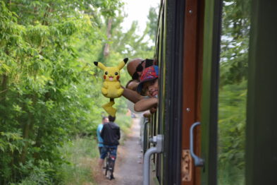 Dzieci wychylają się przez okna stojącego pociągu, trzymając pluszowego Pikachu. Osoba w koszulce i czapce obserwuje z ziemi, opierając się o wagon. Zielone drzewa tworzą tło dla tej kolejowej sceny.