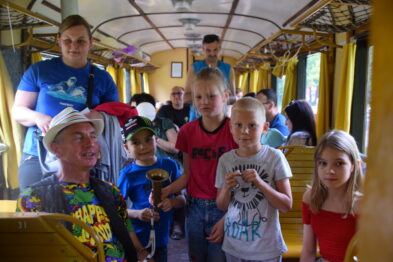 Grupa dzieci wraz z dorosłymi podróżuje w żółtym, drewnianym wagonie kolejowym o tradycyjnym wyglądzie. Jedno z dzieci trzyma w ręku drewniany pociąg, a wszyscy wydają się zainteresowani sytuacją. W tle przez okna widać zieleniące drzewa, co wskazuje na przejażdżkę w dzień.