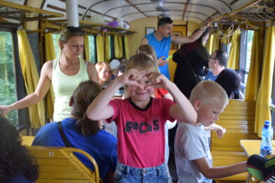 W wagonie wąskotorowym widać grupę ludzi, w tym dzieci i dorosłych podczas jazdy. Dzieci robią różne miny i zabawy, jeden chłopiec na środku tworzy rękami okulary wokół oczu. Wagon ma drewniane siedzenia i okna, przez które wpada światło, co sprawia, że wnętrze jest jasne i słoneczne.