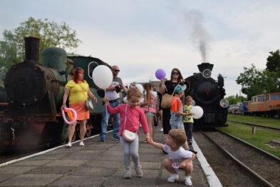 Grupa ludzi, w tym dorośli i dzieci, stoi na peronie w pobliżu starych parowozów. Jedna lokomotywa wydaje parę z dymnicy. Dzieci trzymają balony i inne drobne upominki podczas imprezy kolejowej.
