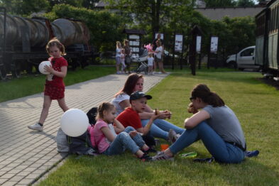 Grupa osób, w tym dorosłych i dzieci, siedzi i odpoczywa na trawniku obok chodnika w muzeum. Niektóre dzieci trzymają białe balony, a tło stanowią eksponaty kolejnictwa, jak wagony. Atmosfera jest swobodna i familijna podczas wydarzenia o tematyce retro kolei wąskotorowej.