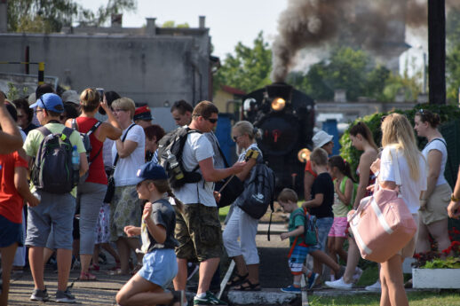 Grupa osób czeka na peronie, w tym dzieci i dorośli wyposażeni w plecaki i torby podróżne, w słoneczny dzień. Czarna parowa lokomotywa zbliża się do peronu emitując kłęby ciemnego dymu. Otoczenie jest czyste i dobrze utrzymane, a podróżni wydają się być w oczekiwaniu na przejażdżkę koleją wąskotorową.