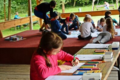 Grupa dzieci siedzi przy drewnianych stołach na zadaszonej platformie i jest zajęta kolorowaniem i rysowaniem. Każde dziecko ma przed sobą kredki, flamastry lub inne przybory do rysowania. W tle widać zielone drzewa, co sugeruje, że aktywność odbywa się na świeżym powietrzu.