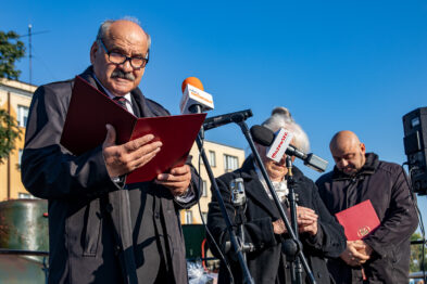 Trzech mężczyzn stoi na zewnątrz podczas jasnego dnia; dwóch z nich trzyma czerwone teczki. Mężczyzna po lewej stronie czyta z kartki, a pozostałe osoby wydają się słuchać; wszyscy noszą odzież oficjalną. W tle widać słoneczne niebo i fragmenty architektury miejskiej.