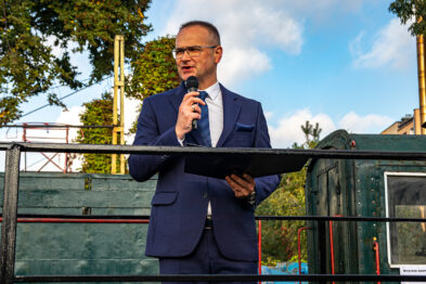 Mężczyzna w ciemnoniebieskim garniturze, białej koszuli i czerwonym krawacie stoi na platformie i przemawia do mikrofonu. Za nim widać zielone drzewa i fragment ciemnozielonego wagonu kolejowego. Mężczyzna trzyma w ręku czarne dokumenty lub folder.
