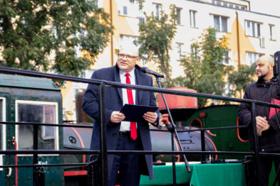 Mężczyzna w garniturze stoi na przemówieniu obok zabytkowej lokomotywy; trzyma kartkę z tekstem. W tle widoczne są drzewa oraz budynek mieszkalny. Obok mężczyzny stoi druga osoba w czarnym ubraniu z mikrofonem.