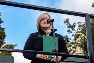 Kobieta stoi na mównicy i przemawia przez mikrofon, trzymając w rękach zielony segregator. Jest ubrana w ciemną bluzę i okulary przeciwsłoneczne. W tle widać niebo i fragmenty roślinności oraz budynku.