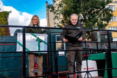 Dwóch mężczyzn stoi na platformie wagonu kolejowego; jeden z nich trzyma mikrofon i dokumenty, podczas gdy drugi obserwuje. Platforma jest ozdobiona zielonymi girlandami i białą wstęgą. W tle widoczne są zabudowania i drzewa, a na pierwszym planie publiczność uważnie słucha przemówienia.