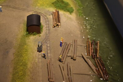 Makieta kolejowa ukazuje fragment torów wraz z przyległym terenem. Widać detalicznie odwzorowane elementy takie jak belki drewniane, figurki ludzi i przydrożne przedmioty. Grupka miniaturowych postaci robotniczych zajęta jest pracą przy układaniu torów.
