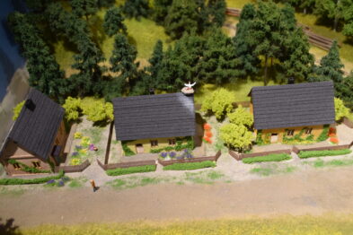 Model wsi z domami pokrytymi ciemnymi dachówkami i otoczonymi kolorowymi ogrodzeniami jest centralnym punktem modelu kolejowego. Otaczają go bogato zielone drzewa i krzewy, podkreślając szczegółowość i realizm makiety. Widać również człowieka w skali miniatury, który dodaje scenie ludzkiego wymiaru.