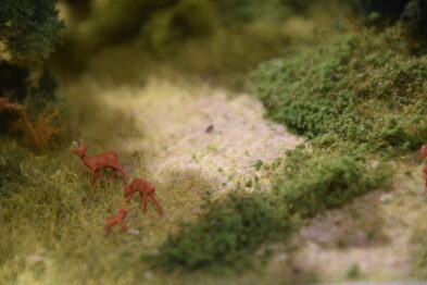 Model terenu pokrytego roślinnością z przewagą niskiej, gęstej zieleni, przedstawia fragment makiety kolejowej. W centralnej części zdjęcia widoczne są trzy miniaturowe modele jeleni na ścieżce otoczonej przez trawę i krzewy. Drobne szczegóły takie jak tekstura roślin i drobina na ścieżce wskazują na wysoki stopień realizmu makiety.