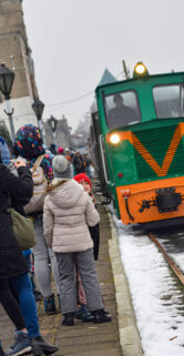 Grupa osób stoi na peronie i obserwuje zielony pociąg wąskotorowy, który podjechał do peronu. Lokomotywa wąskotorowa jest w centrum kadru, a w tle widoczne są inne pojazdy kolejowe oraz budynki stacyjne. Śnieg pokrywa ziemię oraz dachy pociągów, natomiast niebo jest pochmurne.