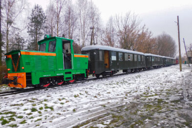 Zielona lokomotywa wąskotorowa stoi na torach obok niej wykładany jest ciemniejszy, pasażerski wagon. Po obu stronach torów widać warstwę śniegu pokrywającą trawę oraz ziemie, a w tle drzewa bez liści. Niebo jest zachmurzone, co sugeruje zimową pogodę.