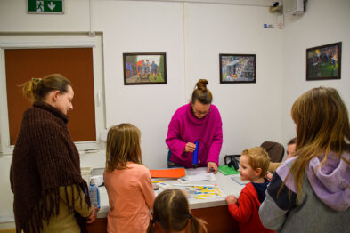 Grupa dzieci wraz z opiekunami uczestniczy w zajęciach prowadzonych na stole przez przewodniczącą osobę. Dzieci są skupione na wykonywaniu zadania, używając papierów i innych materiałów. Sala jest jasna, na ścianach widać kolorowe obrazy z motywami kolejowymi.
