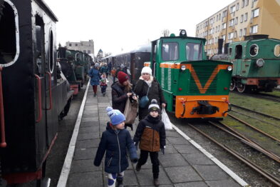 Dorośli i dzieci w zimowej odzieży spacerują obok zielonej lokomotywy oraz ciemnoczerwonych wagonów stojących na torach wąskotorowej kolejki. Na tle niebieskiego nieba i pobliskich budynków, przejrzysty dzień sprzyja wydarzeniu. Zainteresowanie uczestników skierowane jest na pociągi, które są atrakcją imprezy.
