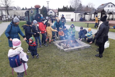 Grupa dzieci i kilku dorosłych gromadzi się wokół otwartego paleniska, na którym płonie ogień. Na tle zimowego krajobrazu widać również zieloną lokomotywę parową i wagony. Wszyscy uczestnicy noszą ciepłe ubrania, a niektóre dzieci mają na głowach kolorowe czapki.