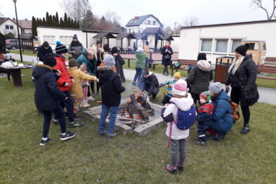 Grupa osób stoi wokół otwartego ogniska, nad którym przygotowywane są pieczone kiełbaski. W tle widać domy i drzewa bez liści, co sugeruje, że jest to zimowa pora. Uczestnicy, wśród których są dorośli i dzieci, ubrani są w ciepłe kurtki i czapki.