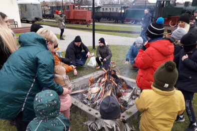 Grupa dzieci w towarzystwie dorosłych stoi wokół ogniska; niektóre dzieci trzymają patyki, a za nimi widać czerwony wagon kolejowy. Osoby mają na sobie zimowe ubrania, takie jak kurtki i czapki. Scena rozgrywa się na otwartej przestrzeni, prawdopodobnie na terenie związanym z kolejnictwem.