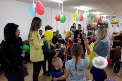 Grupa dzieci i kilku dorosłych uczestniczy w zabawie w jasnym pomieszczeniu, które jest udekorowane kolorowymi balonami. Dzieci są w ruchu i niektóre trzymają balony; jedna z dorosłych osób prowadzi zajęcia. Wszystkie osoby są ubrane w codzienne ubrania, a atmosfera wydarzenia wydaje się być wesoła.