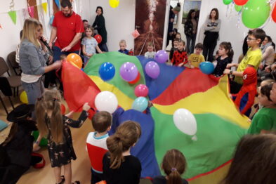 Grupa dzieci wraz z kilkoma dorosłymi bawi się kolorowym spadochronem w pomieszczeniu, gdzie na ścianach widać dekoracje oraz balony. Dzieci są ubrane w kolorowe stroje i trzymają spadochron, pod którym znajdują się kolorowe balony. W tle widać innych uczestników imprezy i dekoracje, tworząc radosną atmosferę.