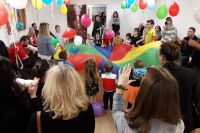Dzieci i dorośli uczestniczą w zabawie z użyciem wielobarwnego spadochronu rozpiętego powyżej ich głów w pomieszczeniu ozdobionym kolorowymi balonami. Przy bocznej ścianie widoczna jest grupa obserwatorów dokumentujących wydarzenie za pomocą smartfonów. Przez całe pomieszczenie unosi się atmosfera świętowania i radosnego wspólnego spędzania czasu.